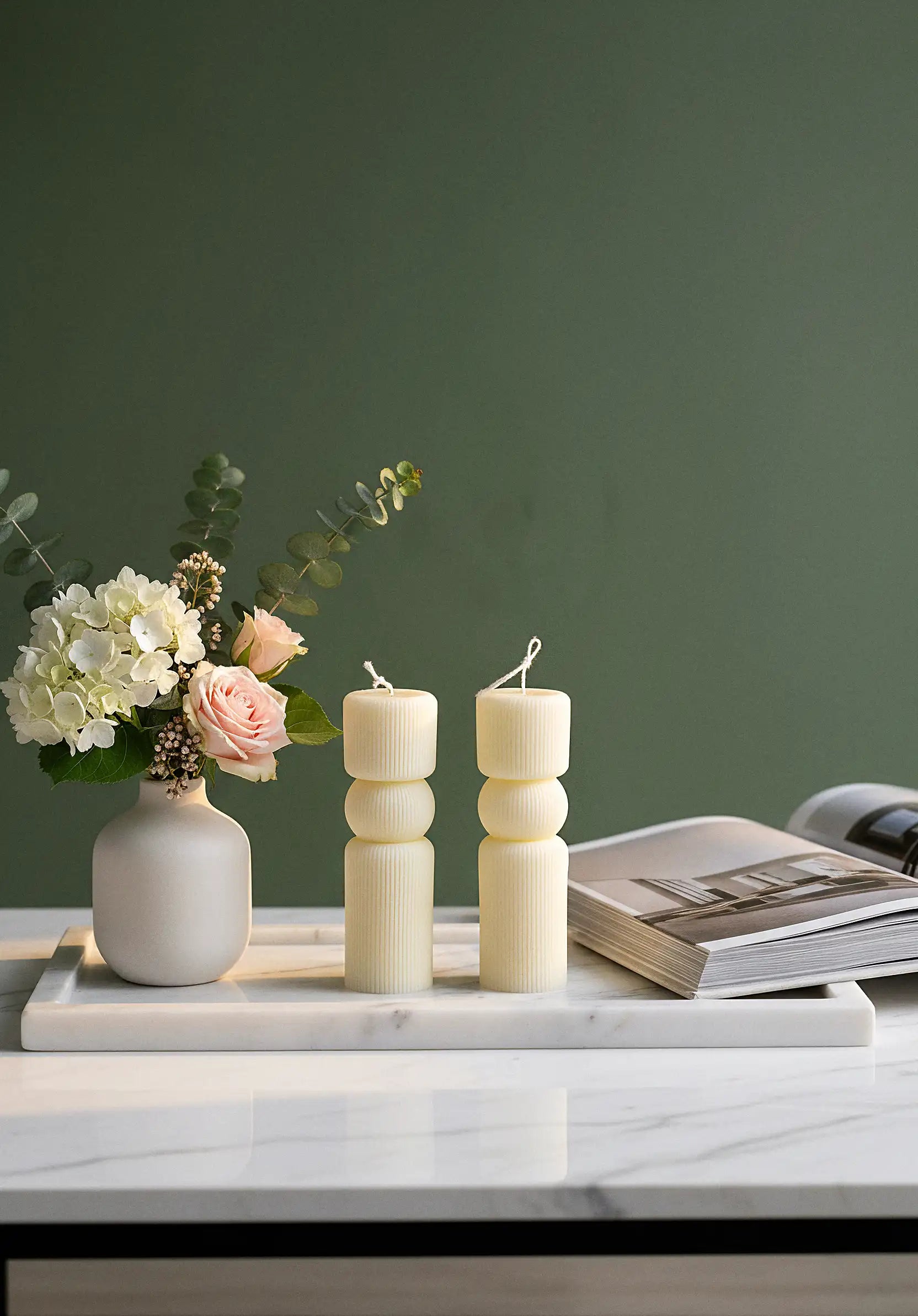 Two white pillar  candles and a vase with flowers on a marble surface against a green wall.